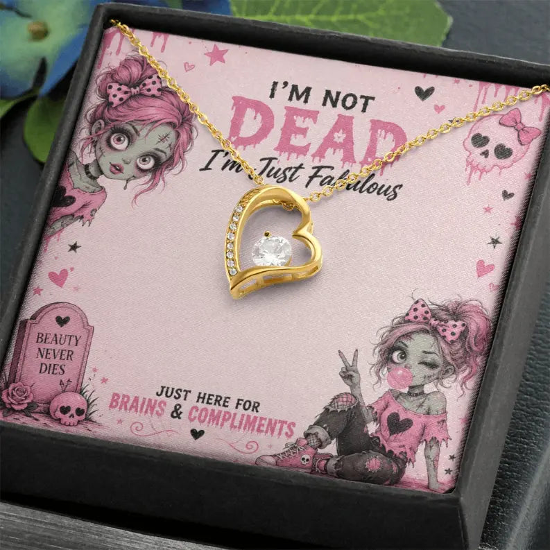 A gold heart-shaped pendant necklace with a clear rhinestone center, displayed on a pink gothic-themed gift card inside a black box. The card features cartoon zombie illustrations and the text 'I'M NOT DEAD I'm Just Fabulous' and 'JUST HERE FOR BRAINS & COMPLIMENTS'.