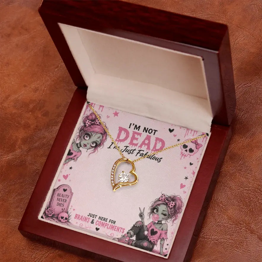 A gold heart-shaped pendant necklace with a clear rhinestone center, displayed inside an open dark wood LED spotlight gift box on a pink printed card with zombie illustrations and the text 'I'm NOT DEAD I'm Just Fabulous'.