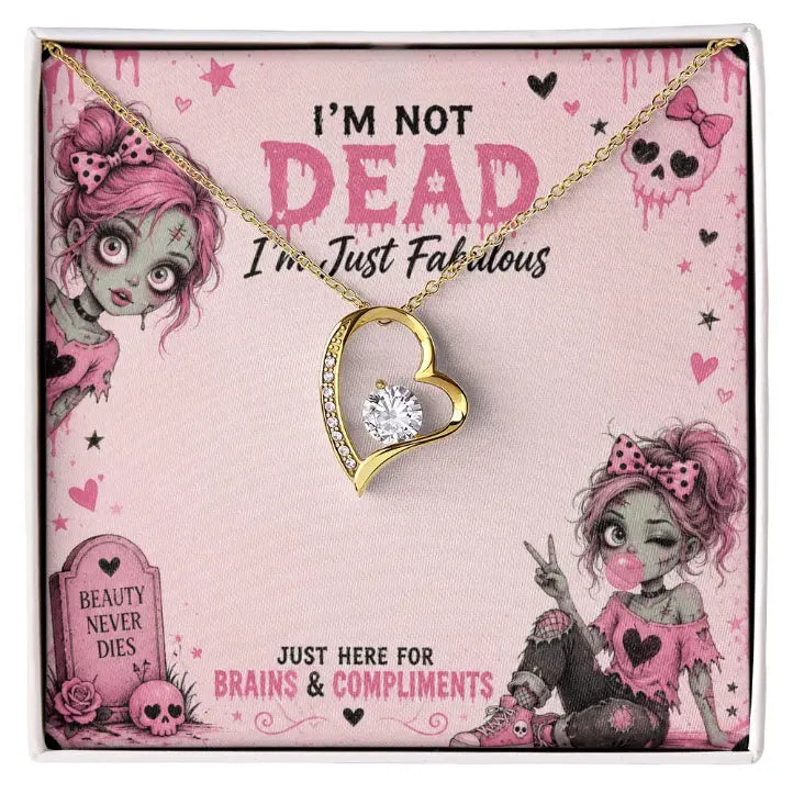 Gold heart-shaped pendant necklace with a clear rhinestone center, displayed on a pink illustrated gift card that reads 'I'M NOT DEAD I'm Just Fabulous' with cartoon zombie and gory girl artwork.