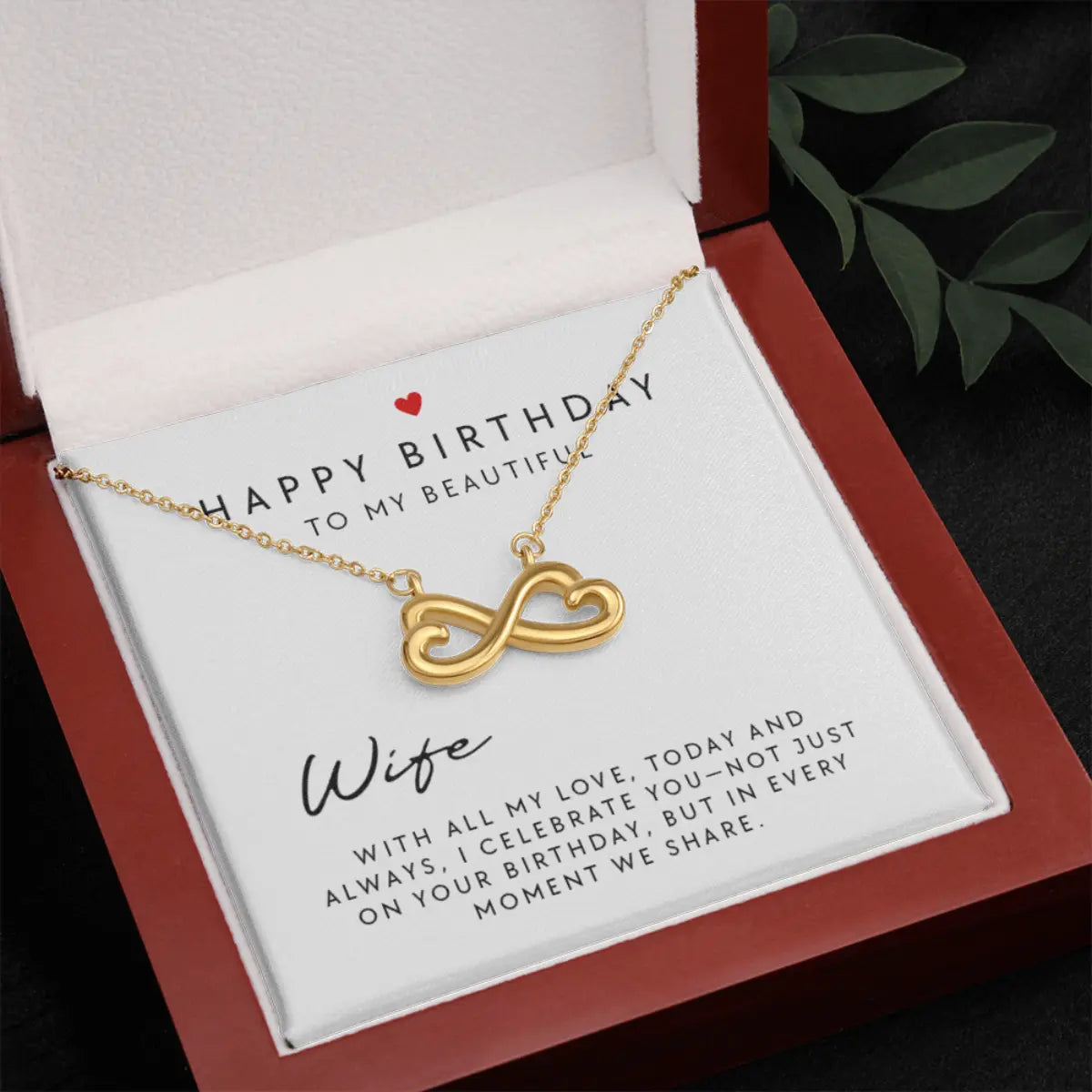 A gold infinity heart necklace presented in a red gift box with a white insert card addressed to a wife, with a dark textured surface and green leaves in the background.