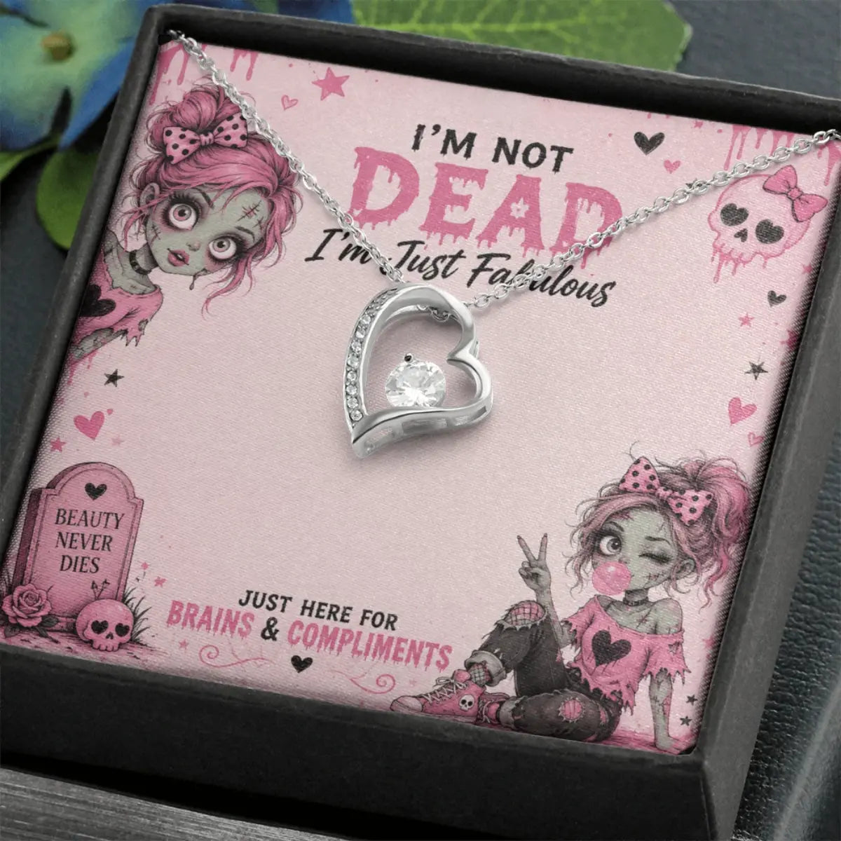 A gothic-themed necklace with a silver heart pendant is presented in a black gift box on a pink card with zombie illustrations and the text 'I'M NOT DEAD I'm Just Fabulous'.