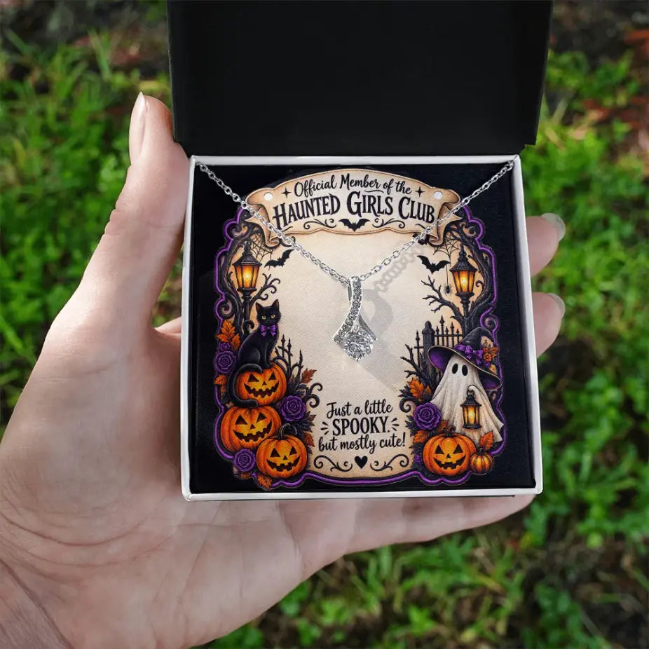 A hand holds an open black gift box containing a silver necklace displayed on a decorative Halloween-themed card. The card reads 'Official Member of the Haunted Girls Club' and features pumpkins, a ghost, and spooky illustrations.