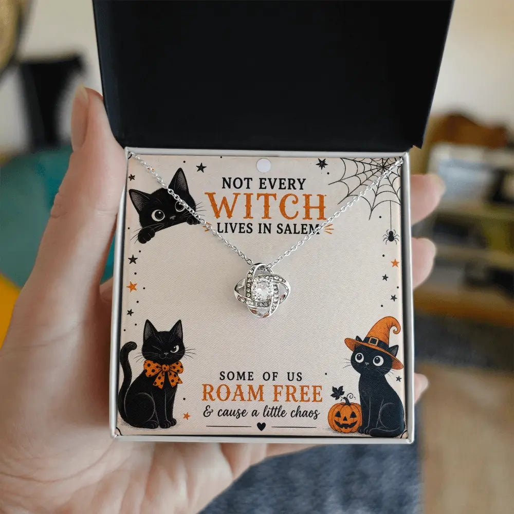 A hand holds an open black gift box displaying a silver necklace with a clear crystal pendant on a printed card. The card features Halloween-themed black cat illustrations and the text 'NOT EVERY WITCH LIVES IN SALEM' and 'SOME OF US ROAM FREE & cause a little chaos'.