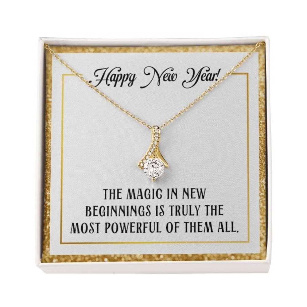 A gold-colored pendant necklace featuring a large, round-cut central crystal dangling from a ribbon-shaped bail encrusted with smaller crystals. The necklace is displayed inside a square white gift box with a sparkling gold glitter border. 
