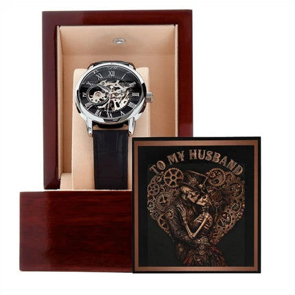 A luxury skeleton wristwatch with an intricate mechanical movement, black outer dial, and black leather strap, displayed inside an open dark wood presentation box. Resting next to the watch is an ornate black-and-copper greeting card that reads "TO MY HUSBAND" in a stylized font, featuring a gothic steampunk illustration of a skeleton husband and wife embracing in love.