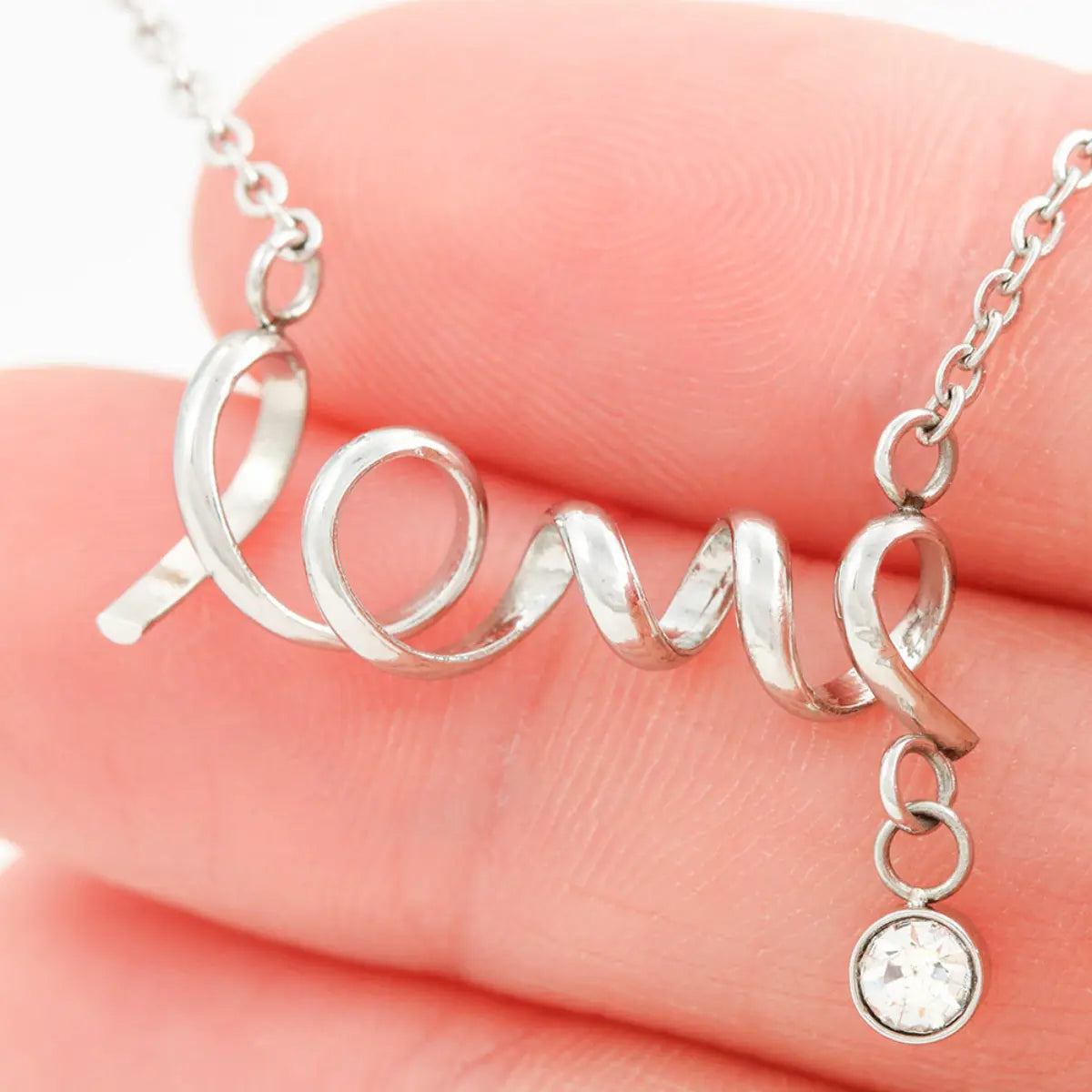 A silver script pendant spelling 'LOVE' draped across a person's fingers, with a small clear round stone charm hanging from one end and part of the silver chain visible at the top.