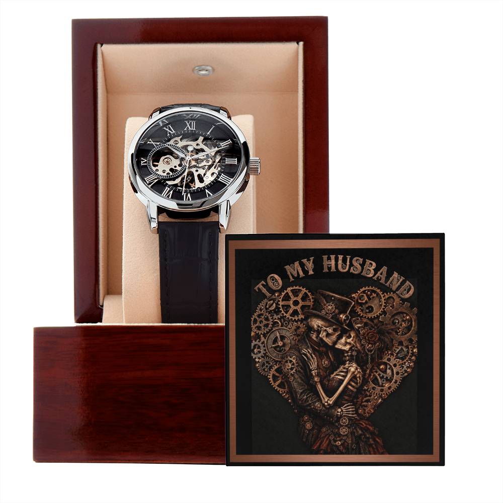 A luxury skeleton wristwatch with an intricate mechanical movement, black outer dial, and black leather strap, displayed inside an open dark wood presentation box. Resting next to the watch is an ornate black-and-copper greeting card that reads "TO MY HUSBAND" in a stylized font, featuring a gothic steampunk illustration of a skeleton husband and wife embracing in love.