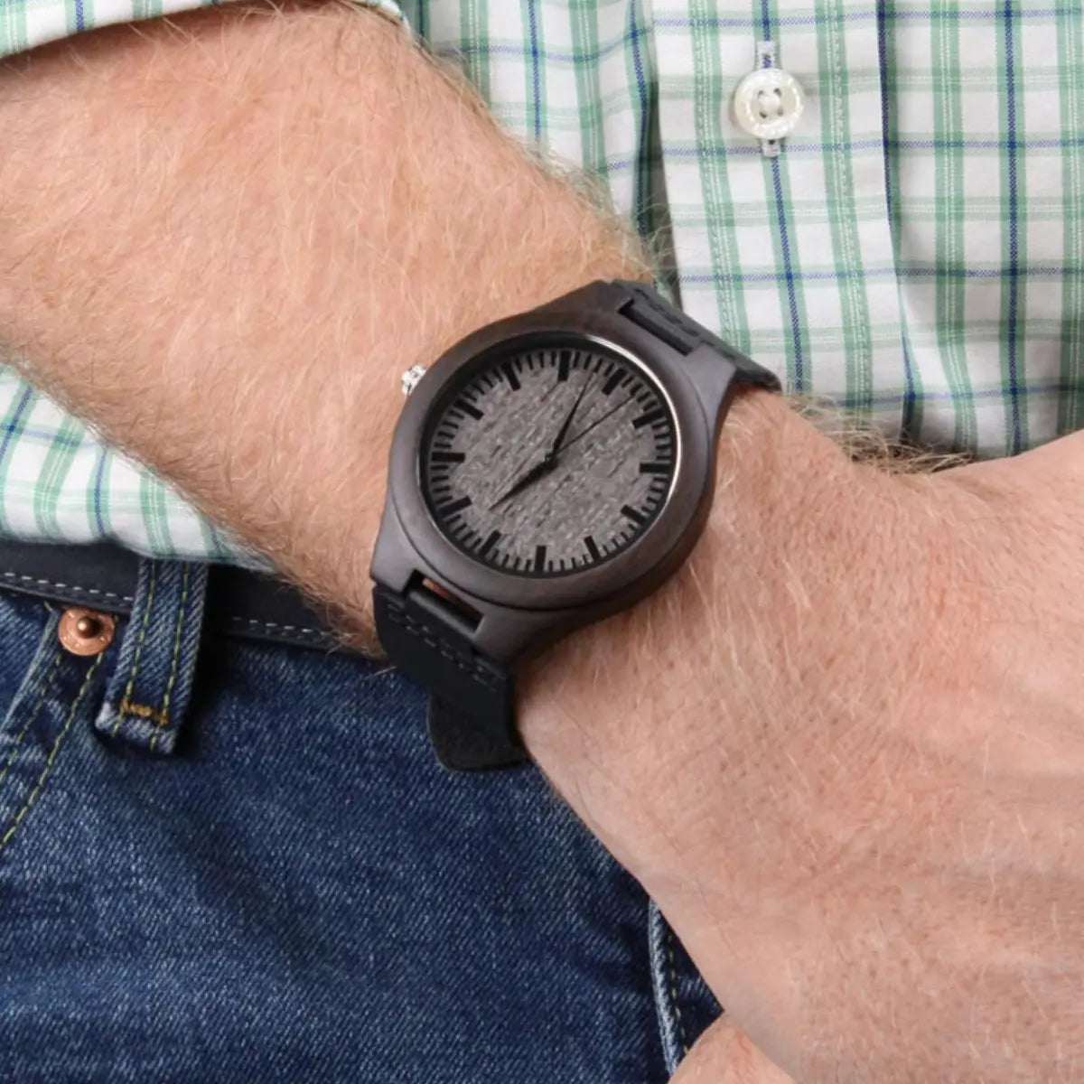 A close-up of a person's wrist wearing a dark brown wooden watch with a textured gray dial and black strap, paired with a green and white plaid shirt and blue jeans.