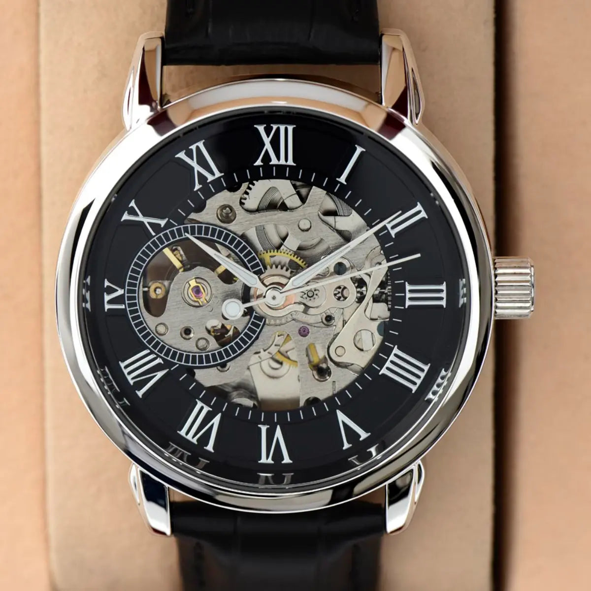 A close-up photograph of a men's openwork skeleton mechanical watch with a silver stainless steel case, Roman numerals on a black outer dial, an exposed silver and gold internal gear movement, and a black textured leather strap against a light beige background.