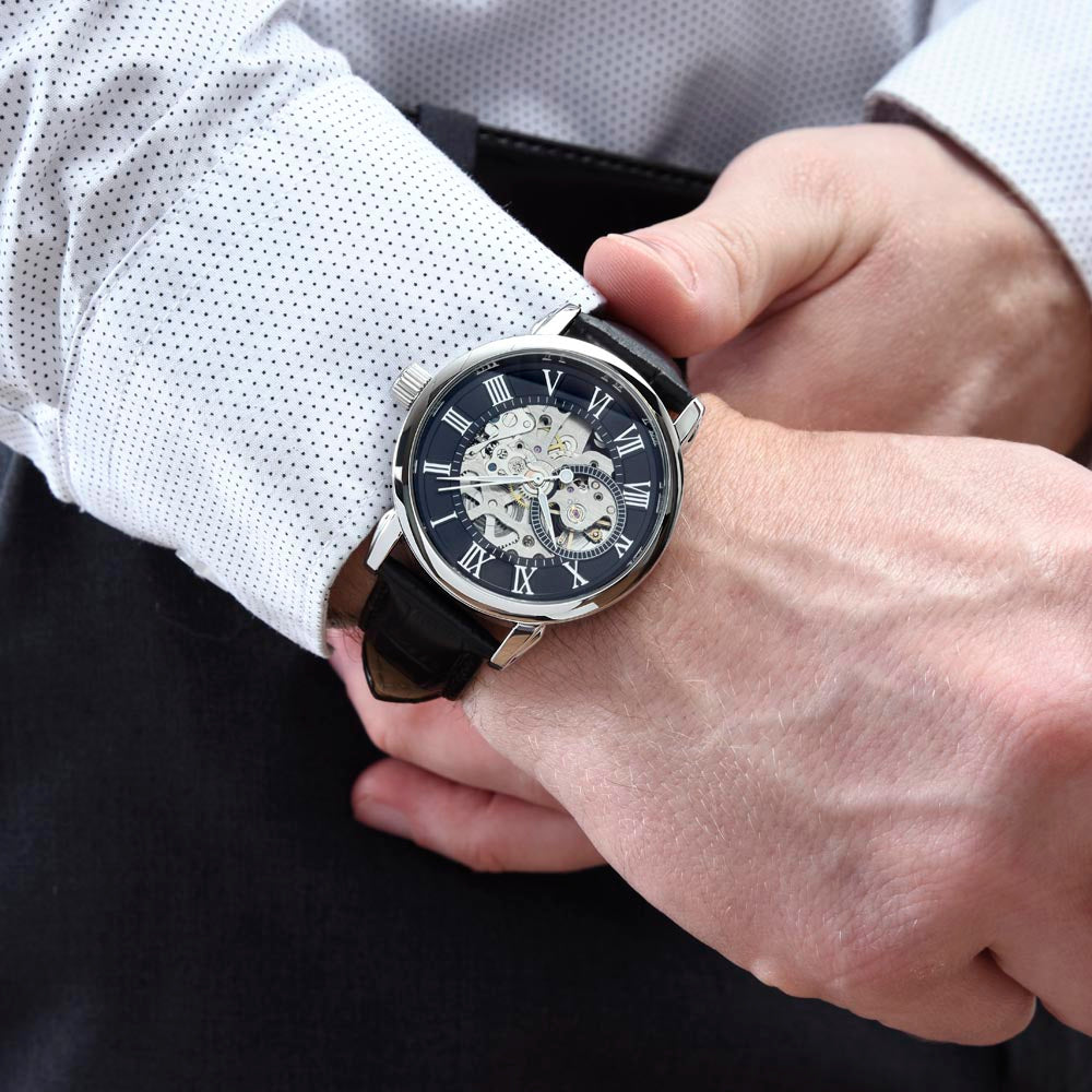 Lifestyle photograph of luxury men's openwork skeleton watch with a black leather strap on a man's wrist, styled with a crisp white dress shirt and dark tailored dress slacks.
