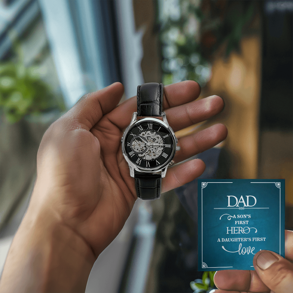 A close-up lifestyle photograph of a man holding a luxury openwork mechanical skeleton watch in his left hand and a personalized message gift card in his right hand. The background features a softly lit room with a large window, warm wooden furniture, and green houseplants.