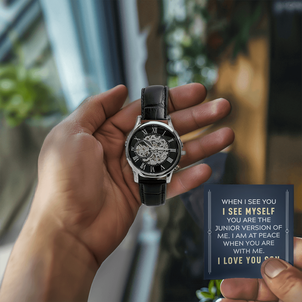 A close-up lifestyle photograph of a man holding a luxury openwork mechanical skeleton watch in his left hand and a personalized message gift card in his right hand. The background features a softly lit room with a large window, warm wooden furniture, and  vibrant green houseplants.