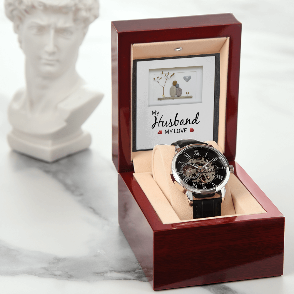 Men's openwork skeleton automatic watch with a genuine leather strap, displayed inside an illuminated luxury mahogany gift box with a soft LED spotlight shining onto a sentimental card for a husband. The background features a classical white powdered wig bust statue on a wooden surface.