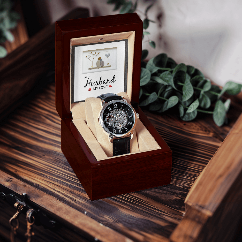 An open mahogany luxury gift box containing a men's openwork skeleton automatic watch and a sentimental husband message card, sitting on a weathered rustic wood table. The background features a softly blurred green houseplant leaf.