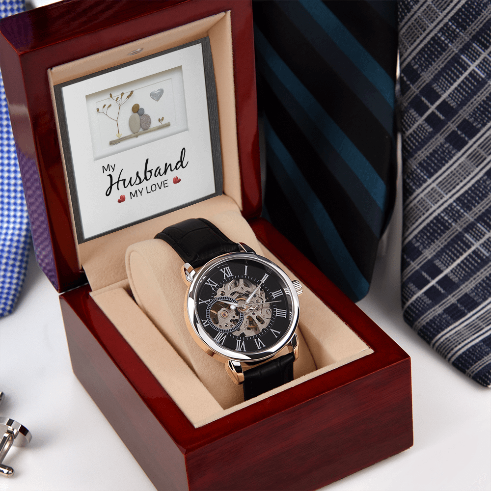 A right-skewed, angled view of a luxury men's openwork skeleton automatic watch with a genuine leather strap, resting inside an open mahogany gift box alongside a sentimental message card. The background displays a neat row of assorted colorful men's ties on a wooden surface.