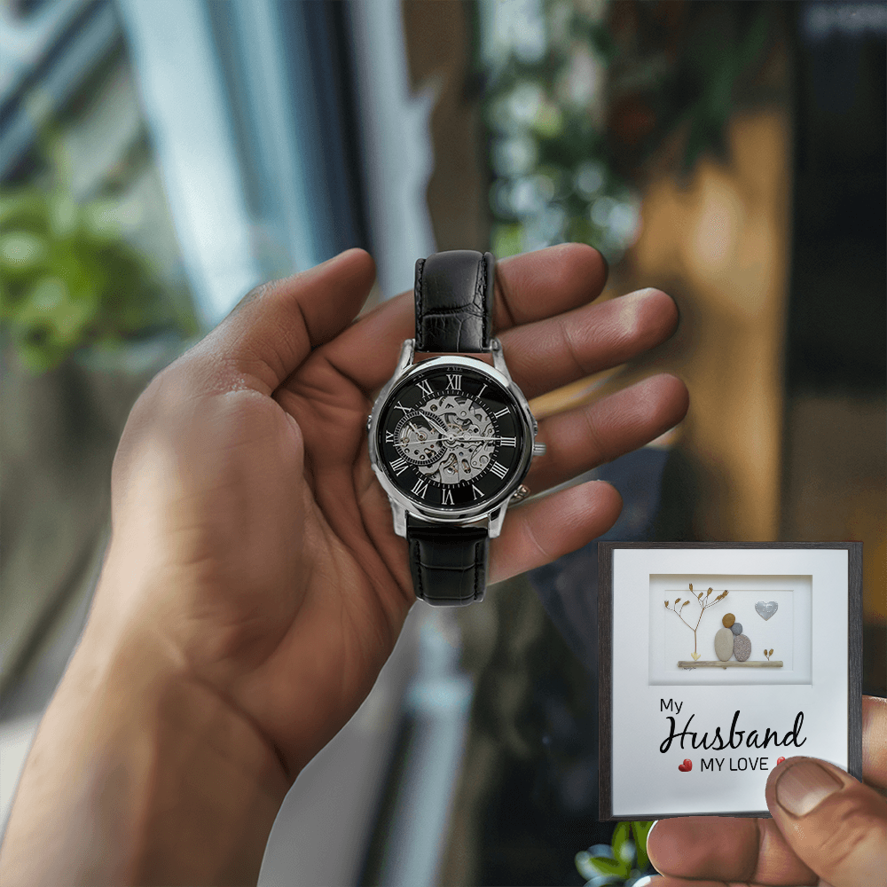 A close-up lifestyle shot of a man holding a luxury openwork mechanical skeleton watch in his right hand and a sentimental husband message card in his left hand. The background features a brightly lit room with a window, warm wooden furniture, and vibrant green houseplants.