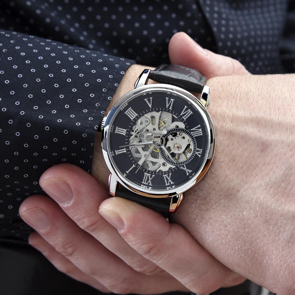 A close-up lifestyle photograph of a men's openwork mechanical skeleton watch with a genuine leather strap being worn on a man's wrist. The man is wearing a tailored dark business jacket with small white dots over a crisp white dress shirt.
