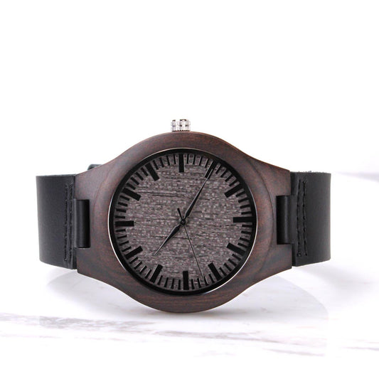 A men's minimalist wristwatch featuring a dark brown wooden bezel and a textured grey wood-grain dial face. The watch has black tick mark hour indices, thin black hands, a silver adjustment crown at the top, and thick black leather straps on a white marble surface.