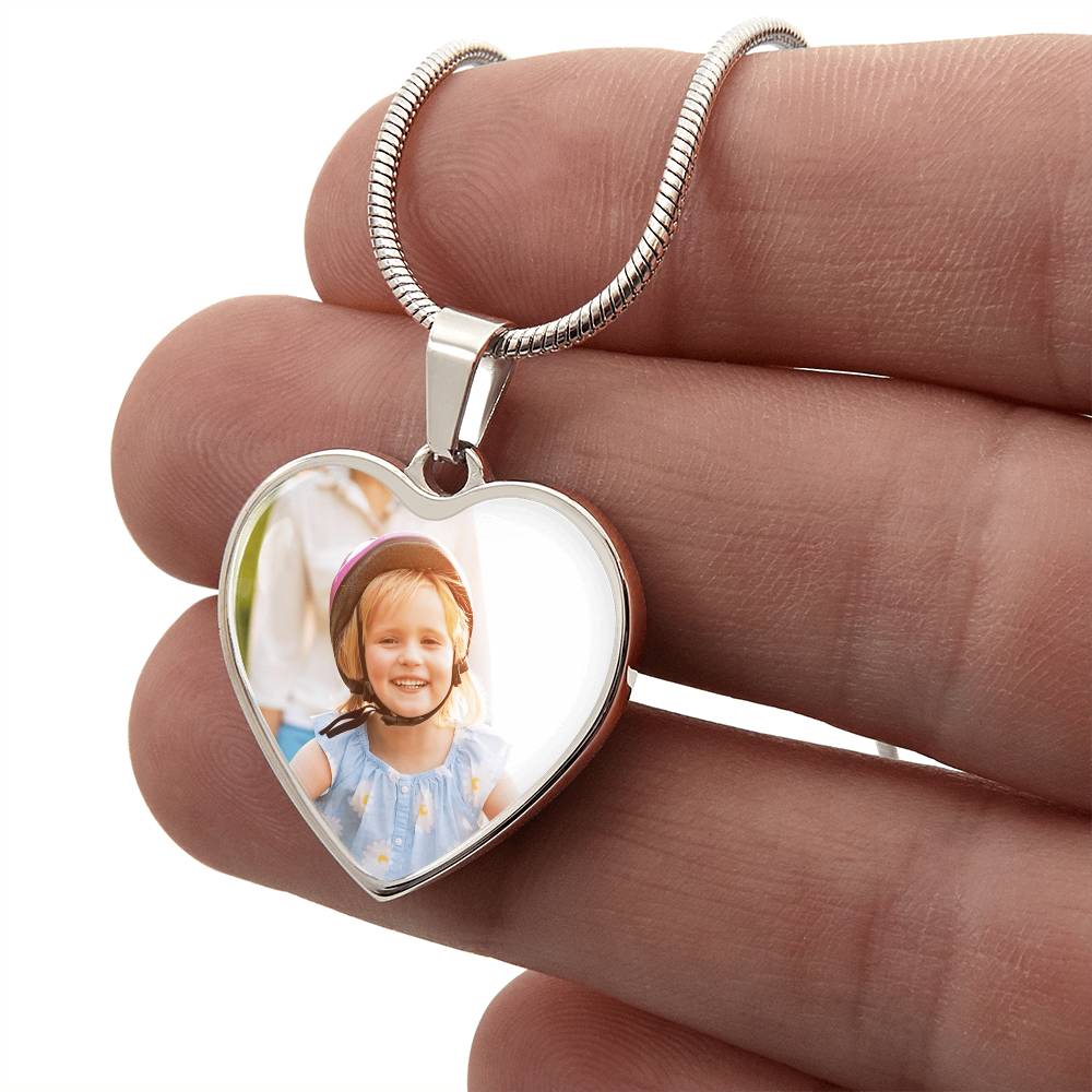 A hand holding a personalized silver heart pendant necklace featuring a custom color photo print of a young girl wearing a helmet, showcasing the detailed scale and craftsmanship of the customized jewelry gift.