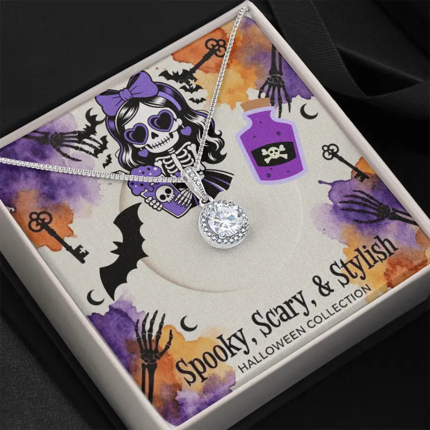 A silver-toned necklace with a clear crystal pendant is displayed inside a Halloween-themed gift box. The box features a printed insert with purple watercolor splashes, black bat silhouettes, skeleton hands, keys, and a cartoon skeleton wearing a purple bow.