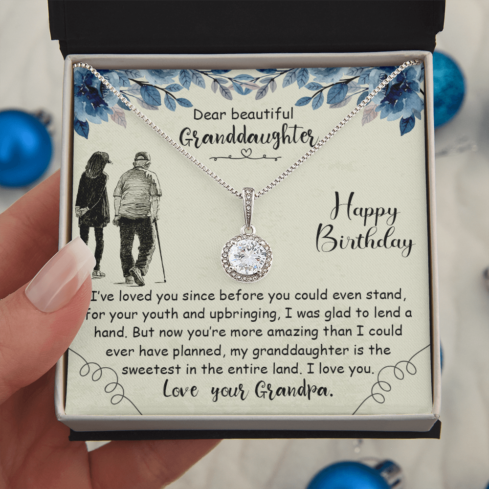 A hand holding a personalized necklace gift card for a granddaughter, featuring a silver pendant with a clear round gemstone on a cream-colored card with blue floral illustrations and a touching birthday message.