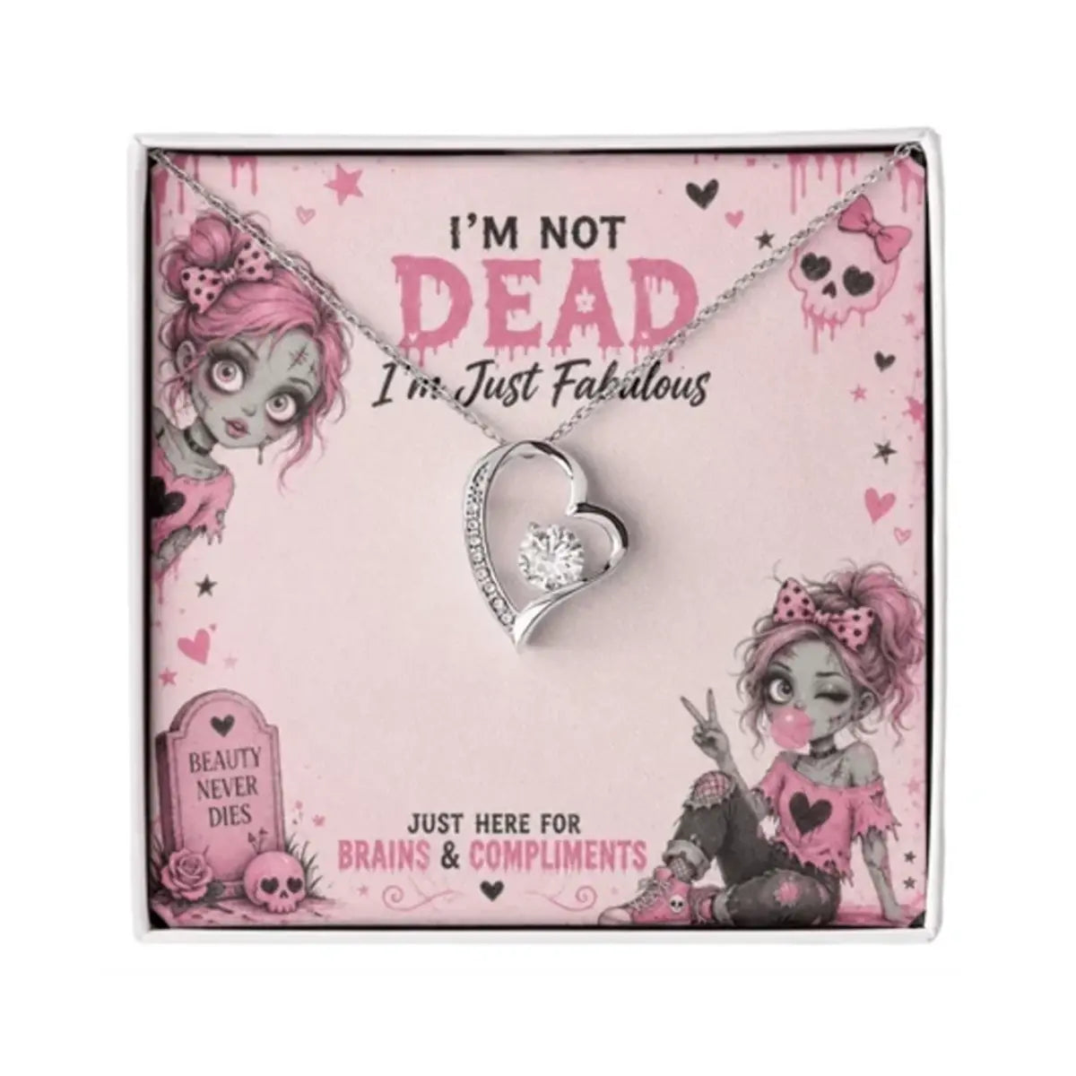 A silver heart-shaped pendant necklace with a clear crystal cubic zirconia center, presented on a pink gothic-themed gift card that reads 'I'M NOT DEAD I'm Just Fabulous'. The card features zombie illustrations and is shown inside its clear outer box against a white background.