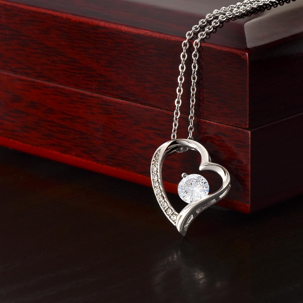 Close-up product photo of a silver heart pendant necklace with an intertwined heart design, sparkling crystal accents, and a large round center stone displayed on a dark red wood box.