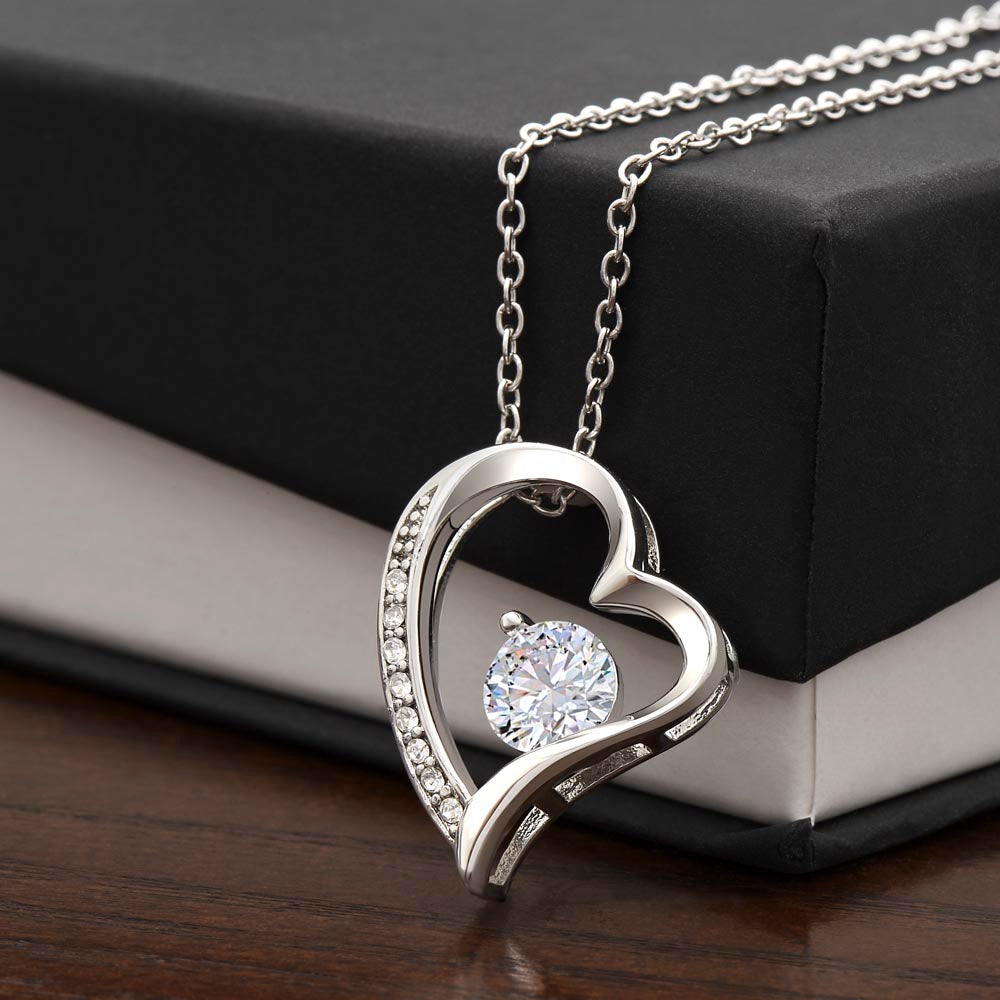 Close-up product photo of a silver heart pendant necklace with an intertwined heart design, sparkling crystal accents, and a large round center stone displayed on a black jewelry box.