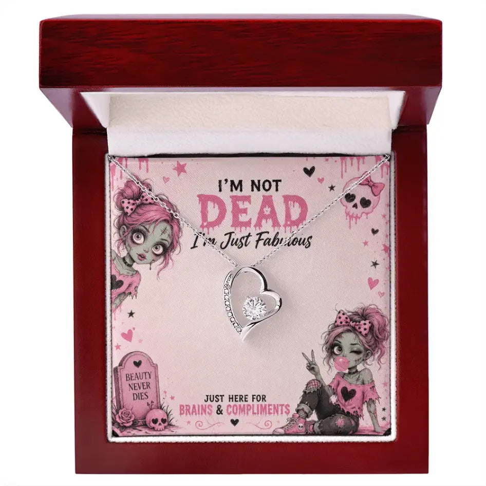 A silver heart pendant necklace with a clear rhinestone is displayed inside an open wooden gift box with LED spotlight. The necklace is attached to a pink printed card with gothic zombie illustrations and the text 'I'M NOT DEAD I'm Just Fabulous'.