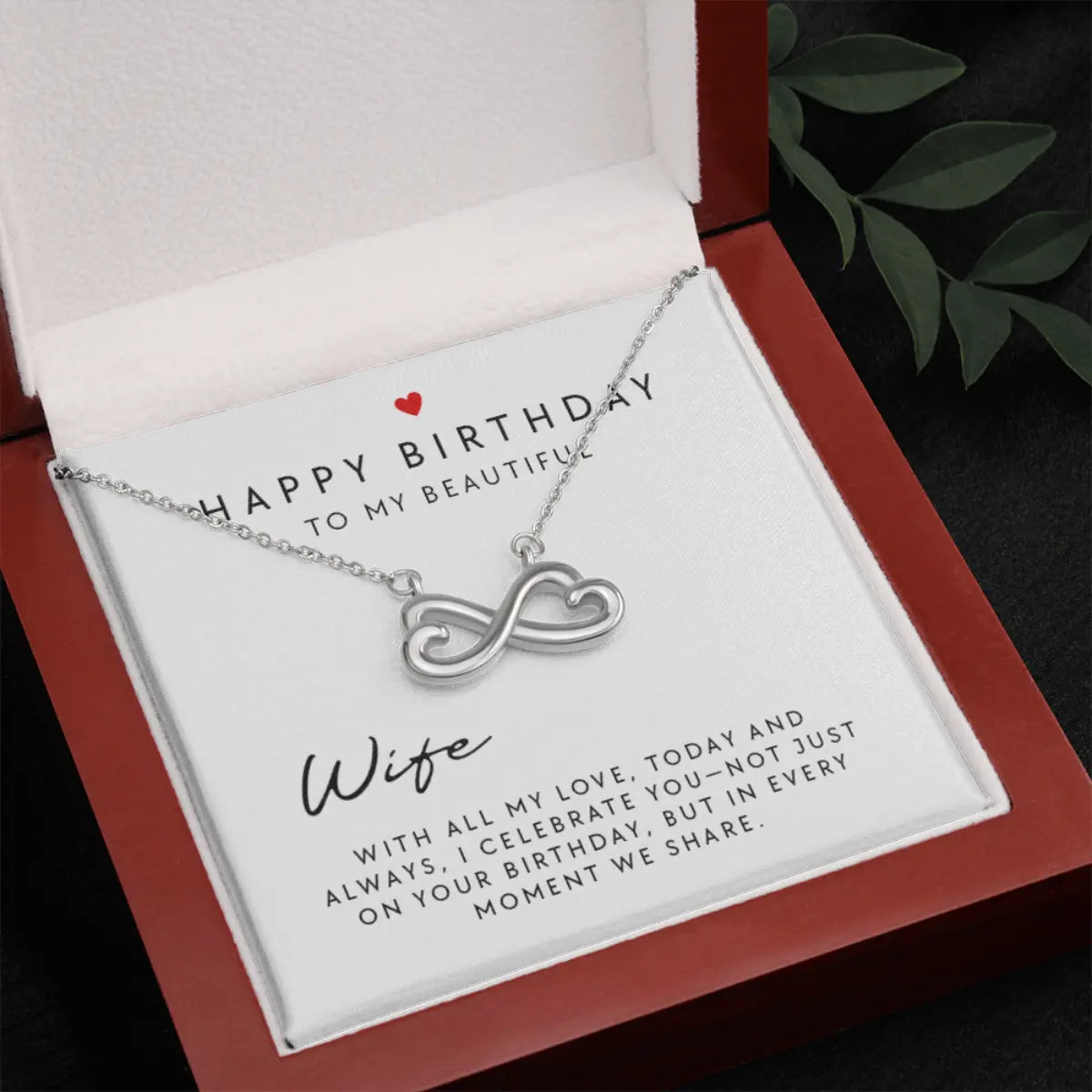 A silver infinity necklace presented in a red gift box with a white card that reads 'Happy Birthday to my beautiful Wife' and a sentimental message. The box is photographed on a dark surface with a leafy branch in the corner.