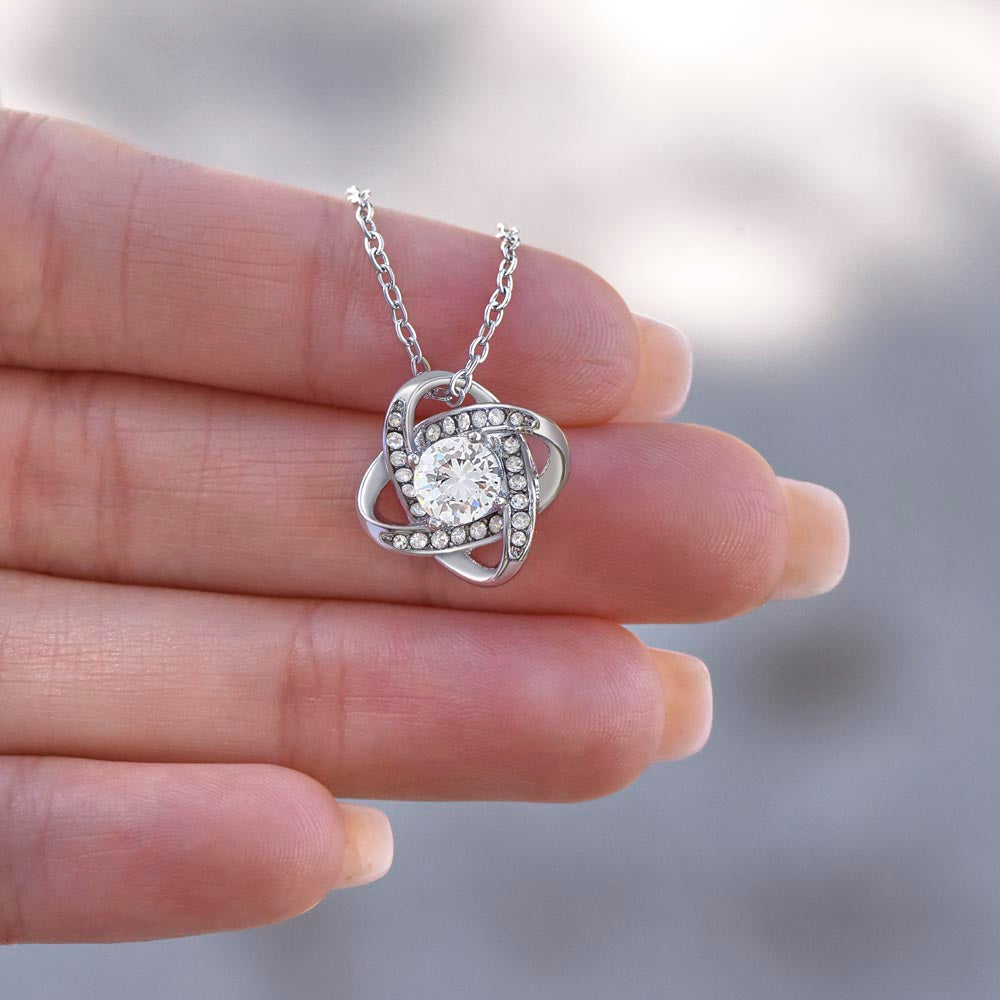 A silver-toned necklace with a knot-shaped cubic zirconia pendant, presented on a woman's open hand.