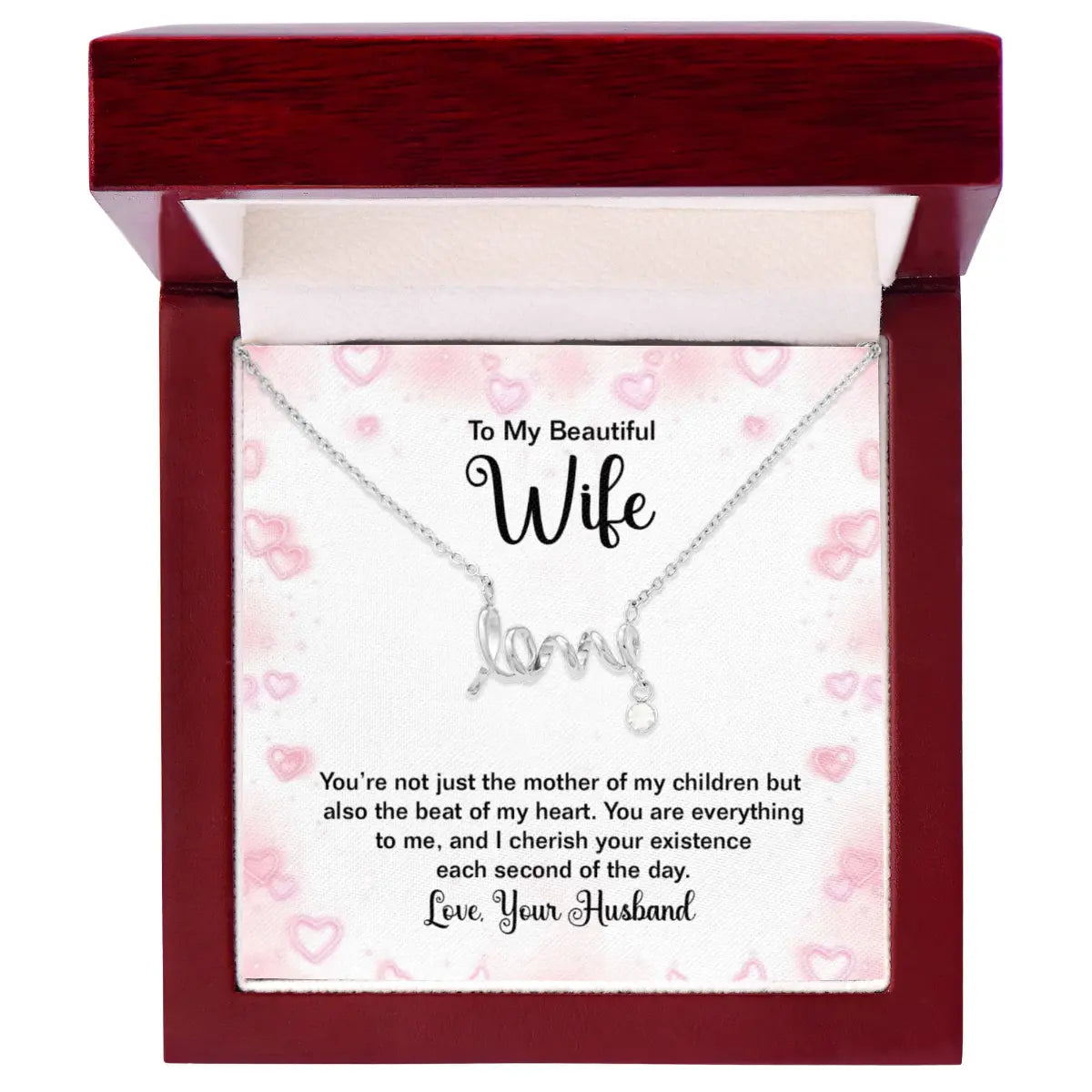 A silver necklace with a 'love' pendant is presented in an open dark red wooden gift box. The necklace is displayed on a white card with pink hearts and sentimental text addressed to a wife, with 'Love, Your Husband' written at the bottom.