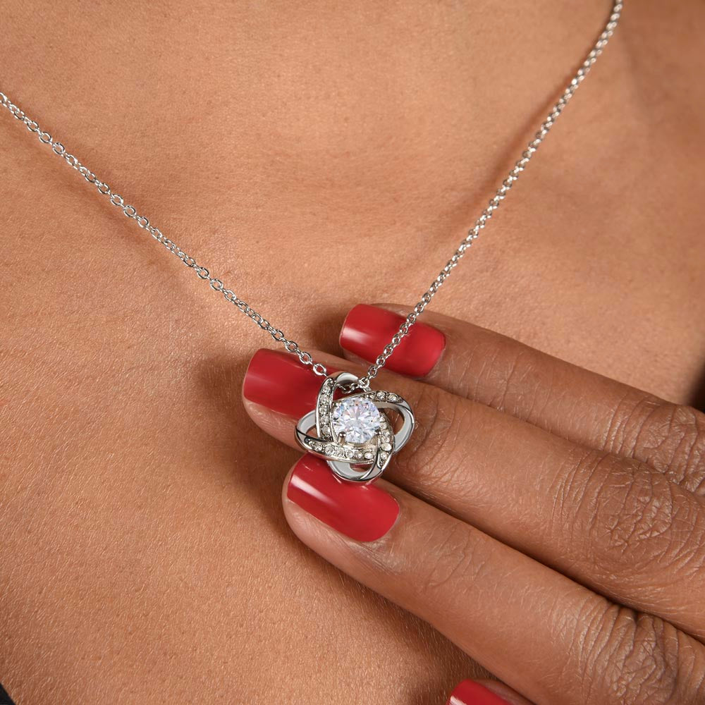 A close-up shot of a person's hand with bright red nail polish gently lifting a silver love knot pendant necklace. The pendant features an intertwined knot design encrusted with small pavé crystals surrounding a brilliant round-cut center gemstone, resting against the skin.