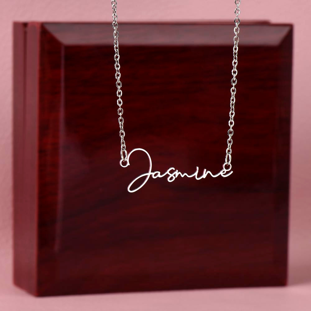 Silver personalized signature style name pendant necklace draped over a dark red gift box against a solid pink background.