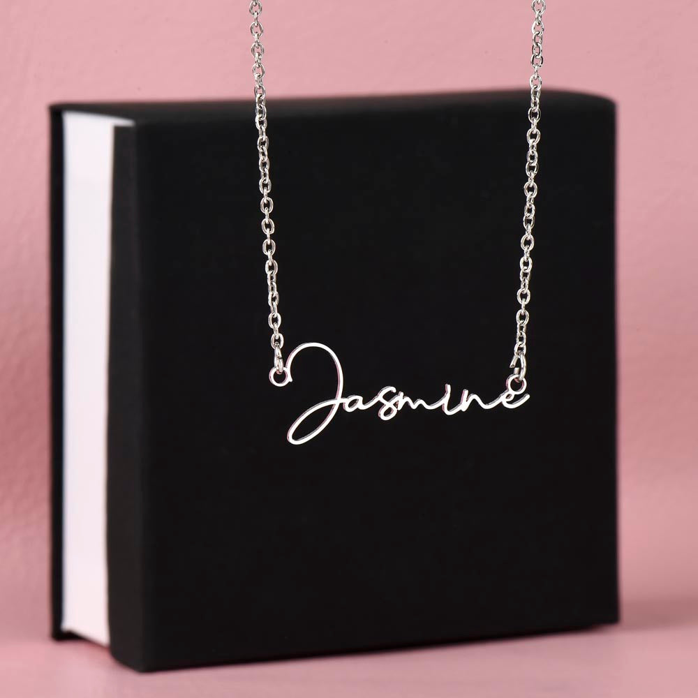 Silver personalized signature style name pendant necklace draped over a black and white gift box against a pink background.