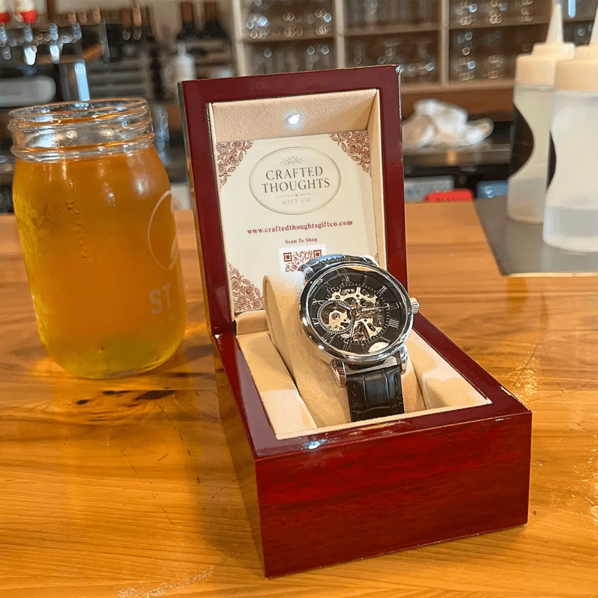 A skeleton wristwatch with a black dial and dark strap displayed inside an open reddish-brown wooden presentation box on a wooden table. A glass jar filled with amber liquid sits beside it, and the background shows a softly blurred bar or cafe interior.