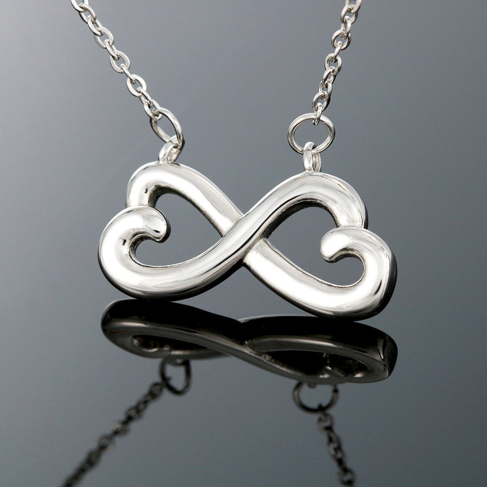 a silver endless love infinity heart necklace suspended and mirrored by a black mirror below.